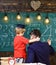 Young male teacher guides his child student to learning while boy hugs teacher, sitting in classroom, chalkboard with