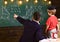 Young male teacher guides his child student to learning while pointing and looking at chalkboard with scribbles on