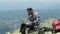 Young male traveler working on laptop outdoors on top of mountain.