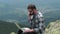 Young male traveler working on laptop outdoors on top of mountain.