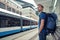 Young male traveller with backpack and smartphone stand on public transport stop and waiting tram in modern city center