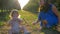 Young mama gives glass of clean water to small daughter sitting on grass between rows of trees on nature during harvest