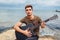 Young man with acoustic guitar playing on beach surrounded with rocks on rainy day