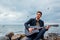 Young man with acoustic guitar playing and singing on beach surrounded with rocks on rainy day