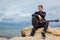 Young man with acoustic guitar sitting on beach surrounded with rocks on rainy day