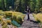 Young man with a backpack and tripod is walking along the bridge across a mountain stream into the depths of the forest towards ad