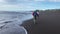 Young man with a backpack walks along the black sand beach and runs away from the rolling waves.