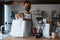 Young man barista in face mask and apron giving pos terminal to customer in coffee shop