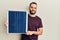 Young man with beard holding photovoltaic solar panel looking positive and happy standing and smiling with a confident smile