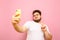 Young man with beard and overweight stands on a pink background, takes a selfie on smartphone camera and shows gesture peace.