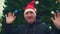 Young man with beard in santa hat on christmas tree background. A happy man dances waving his hands, shows his tongue.