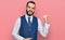 Young man with beard wearing business vest smiling with happy face looking and pointing to the side with thumb up