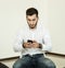 A young man with a beard in a white shirt works with a smartphone while sitting on a chair in a bright room