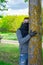 Young man in black medical mask and gloves hugging tree in woodland. Adult male enjoying nature in countryside in period