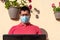 Young man with blue face mask. He is working with his laptop from the balcony of his house.