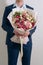 Young man in a blue suit holds in hand a big bouquet of white-pink roses and eustoma in pink packaging on the white wall