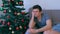 Young man is boring sitting near Christmas tree on couch at home waiting eve.