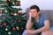 Young man is boring sitting near Christmas tree on couch at home waiting eve.