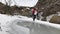 Young man carries his girlfriend on back across puddle covered with melted water.