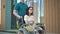 A young man carries a young woman in a wheelchair. An assistant carries a disabled person in a wheelchair along the