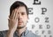 Young man is covering his face with hand and checking his vision. Chart for eye sight testing in background