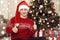 Young man dressed in retro sweater and sanat hat sit near christmas tree. New year eve, ornated fir tree. Winter holiday and love