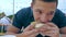 A young man eats a hamburger or cheeseburger with a green bun in an open-air cafe at a festival or village fair.