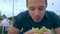 A young man eats a hamburger or cheeseburger with a green bun in an open-air cafe at a festival or village fair.