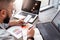 Young man, entrepreneur, freelancer sits in office at table, uses smartphone, working on laptop, makes notes in notebook