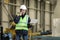 Young man, factory engineer in a work dress and white hard hat holding smartphone and laptop organizing and controlling work