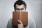 A young man with glasses and a beard covered his face with a book. Education and training. Close-up. Gray background