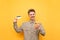 A young man in glasses and shirt stands on a yellow background with a smartphone and a bank card in his hand, looks into the