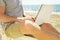 Young man in grey shirt sitting at the beach, working on laptop.