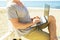 Young man in grey shirt sitting at the beach, working on laptop. Blogger, writer, coder, remote worker surfing browsing researchin