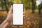 Young man hand holding smartphone with white screen outdoors against autumn sidewalk