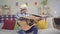 Young man in a hat with a bionic prosthetic arm plays an acoustic guitar sitting on a sofa