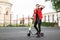 Young man hipster in sneakers in a stylish red denim vest in black jeans in sunglasses stands with an electric scooter