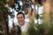 Young man with only his head visible framed between branches of a green tree smiling happily outside in a park. Optimistic and