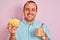 Young man holding bowl with extruded corn standing over isolated pink background happy with big smile doing ok sign, thumb up with
