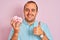 Young man holding bowl with marshmallows standing over isolated pink background happy with big smile doing ok sign, thumb up with