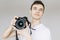 A young man holds a photo camera in his hand and looks straight. Isolated gray background