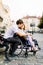 Young man hugs his pretty woman on the wheelchair from behind, while sitting on the bench on the street of old city