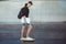 A young man with a laptop under his arm stands on the background of a gray wall, putting one foot on a yellow skate.