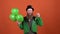 Young man leprechaun celebrating saint patrick`s day isolated on orange wall with balloons