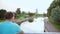 A young man looks at the river standing in the park on the bridge, rear view, camera movement