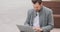 Young man with mustaches and a beard sitting on a bench in the square, typing on the laptop keyboard and speaking to