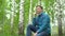 A young man in nature is talking on the phone through headphones. A man sits on a stump in a birch forest with