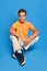 Young man in orange undershirt and white sweatpants sits on the floor over blue background