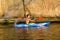 Young Man Paddling Blue Kayak on Beautiful River or Lake under High Rock in the Evening