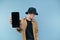 Young man in panama stands on a blue background and shows to the camera a smartphone with a black blank screen, focus on the phone